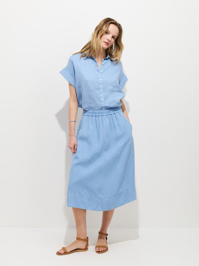 A woman stands against a white background, wearing the garment-dyed Maddie Blouse in light blue (100% linen), tucked into the matching Maddie Skirt In Linen—both in blue—and paired with tan sandals.