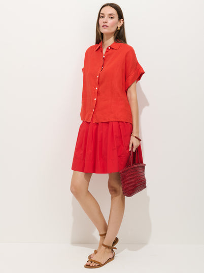 A woman with straight brown hair and a neutral expression stands against a white background wearing the Pippa Skirt In Cotton, a loose red button-up top, brown sandals, and holding a red woven handbag.