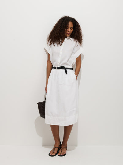 A woman with curly hair stands against a white background, wearing the Maddie Skirt in Linen with a short-sleeve button-up white top, black belt, black sandals, and holding a black woven bag. She looks down with her hands in her pockets.