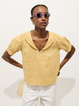 A person wears the Madeleine Shirt In Linen—a yellow short-sleeve linen top with a sailor collar—paired with white pants, purple sunglasses, and hoop earrings, standing hands on hips against a plain background.