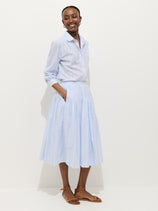 A woman smiles against a white background, wearing a light blue button-up shirt with rolled sleeves and the Hannah Striped Skirt in cotton voile. Brown sandal flats and her hands in her pockets complete the effortless look.