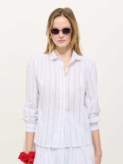 A woman in pink sunglasses wears the Hannah Striped Shirt In Cotton Voile, a white blouse with thin vertical stripes, standing against a plain white background and holding a red flower in her left hand.