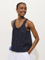 A smiling woman stands against a plain background wearing the Sidney Top in Linen, a relaxed fit, sleeveless navy blue button-up with a bow neckline, paired with a white pleated skirt. Her hair is pulled back and one hand is in her pocket.