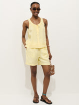 A person wears the Hudson Short In Linen with a sleeveless yellow top and sunglasses, smiling against a white background. They accessorize with a brown woven handbag, black sandals, and a turquoise anklet.
