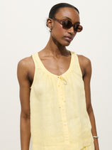 A woman in dark sunglasses wears the Sidney Top In Linen—a sleeveless, light yellow 100% linen button-up with a relaxed fit, scoop neckline, and small bow at the collar—standing against a plain light background.