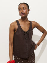 A person stands against a white background, wearing the Sidney Top in Linen—a relaxed fit, sleeveless dark brown button-up with a tie neckline—paired with brown polka dot pants. Their hair is pulled back and they hold a red scrunchie.