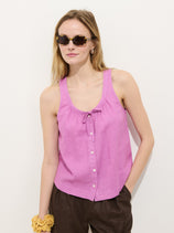A woman stands against a white background wearing the Sidney Top In Linen—a sleeveless pink button-up in a relaxed fit—paired with dark brown pants. She has sunglasses, a yellow scrunchie on her wrist, and her hair down.