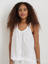A woman with long, curly hair wears the Sidney Top In Linen—a sleeveless, white 100% linen button-up with a round neckline and bow detail in a relaxed fit—standing against a plain white background.