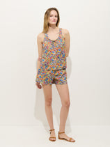 A woman with light skin and straight shoulder-length hair stands against a white background, wearing the Sidney Floral Short In Liberty Fabric—a colorful, relaxed-fit sleeveless romper—paired with brown flat sandals. She appears relaxed.