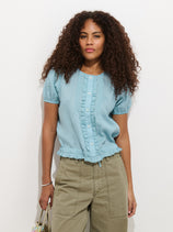 A woman with curly hair wears the Brittany Top In Ramie—a light blue, short-sleeve blouse—paired with olive green high-waisted pants, standing against a plain white background and holding a small bag.