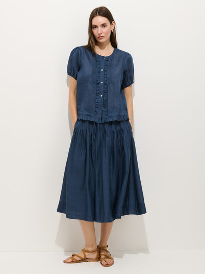 A woman with long brown hair wears the Josephine Skirt in Ramie—a navy blue, fully lined midi skirt with a pull-on waistband and pockets—paired with a matching short-sleeve button-up blouse and tan sandals against a white background.