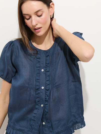 A woman with light skin and straight brown hair wears the Brittany Top In Ramie, a blue short-sleeve blouse with ruffle and button details. She looks down, touching her neck, against a plain light background.