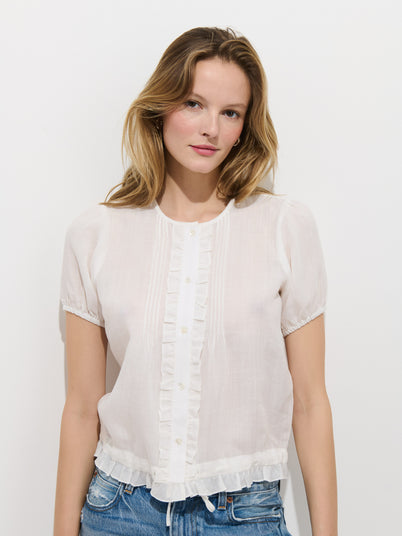 A woman with light brown hair wears the Brittany Top In Ramie, a short-sleeved white blouse with ruffle details, paired with blue jeans. She stands against a plain white background and gazes at the camera with a soft smile.