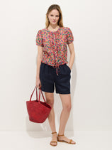 A woman with long blonde hair stands against a white wall wearing the Hudson Short In Linen, a colorful floral blouse, tan sandals, and holding a red woven bag. She looks relaxed and casual, with one hand in her pocket.