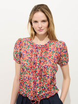 A woman with long, light brown hair wears the Brittany Floral Top in cotton silk, featuring short sleeves and a ruffled drawstring hem, as she poses against a plain white background with a slight smile.