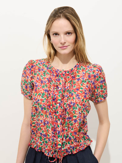 A woman with long, light brown hair wears the Brittany Floral Top in cotton silk, featuring short sleeves and a ruffled drawstring hem, as she poses against a plain white background with a slight smile.