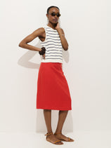 Someone stands against a plain white background wearing sunglasses, a sleeveless white striped top, brown sandals, and the Maya Pencil Skirt in Linen—an elegant red summer skirt falling below the knee.