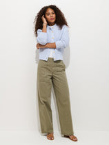 A woman with curly hair wears the Neil Trouser in Herringbone, olive green wide-leg pants, a light blue buttoned cardigan, and tan sandals, standing against a plain white background.
