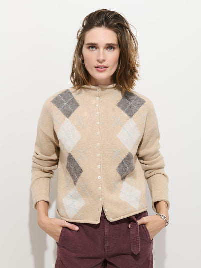 A person with shoulder-length brown hair wears the Taylor Argyle Cardigan in a beige wool-cotton blend and maroon pants, standing against a white background with hands in pockets, looking directly at the camera.
