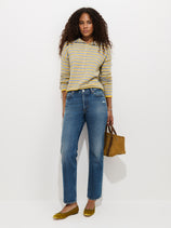 A woman with curly hair wears a gray and yellow striped sweater, the Jane Mid-Rise Straight Jean crafted from 100% cotton denim, and yellow flats. She holds a small brown bag and stands against a plain white background.