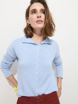 A woman with shoulder-length brown hair wears the Taylor Cardigan in Cotton Cashmere, featuring Troca shell buttons, and rust-colored pants as she stands against a plain white background, looking upward.