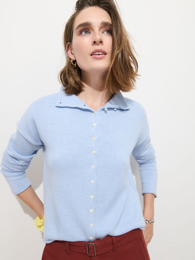 A woman with shoulder-length brown hair wears the Taylor Cardigan in Cotton Cashmere, featuring Troca shell buttons, and rust-colored pants as she stands against a plain white background, looking upward.