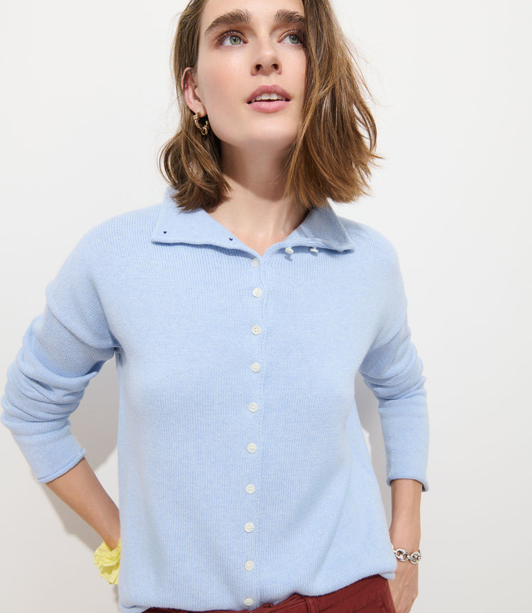 A woman with shoulder-length brown hair wears the Taylor Cardigan in Cotton Cashmere, featuring Troca shell buttons, and rust-colored pants as she stands against a plain white background, looking upward.