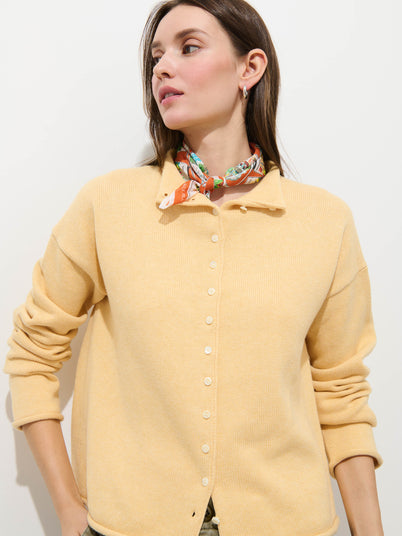 A woman with long brown hair and small hoop earrings looks left, wearing the Taylor Cardigan in Cotton Cashmere (light yellow, oversized boxy fit) and a printed silk scarf, standing against a plain white background.