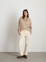 A woman with straight brown hair stands against a white wall, hands in pockets, wearing The Neil Pant In Herringbone with a loose beige sweater and dark loafers for a relaxed, utilitarian look.