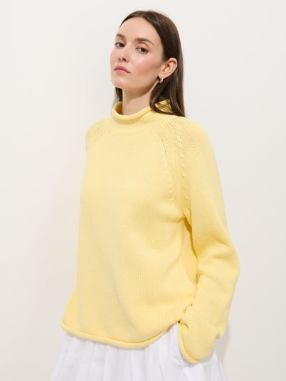 Woman with long brown hair wears the Eastport Rollneck in 100% cotton, yellow, paired with a white skirt. She stands against a plain white background, hands in pockets, gazing slightly upward.