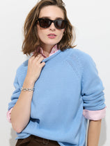 A woman with shoulder-length brown hair and dark sunglasses stands against a plain white background, wearing The Eastport Rollneck In Cotton—a light blue 100% cotton sweater—layered over a pink collared shirt and accessorized with a silver bracelet.