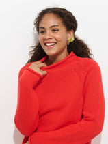 A woman with curly hair smiles brightly, wearing The Eastport Rollneck In Cotton—a vibrant red 100% cotton sweater—and gold hoop earrings, standing against a plain white background.