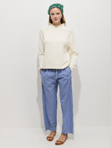 A woman stands against a white background wearing the Hudson Striped Pant In Poplin—100% cotton blue and white wide leg pull-ons—with a cream knit sweater, brown sandals, and a green patterned headscarf, hands in her pockets.