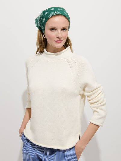 A woman wearing the Eastport Rollneck In Cotton, blue and white striped pants, and a green bandana with white patterns stands against a plain white background with her hands in her pockets.