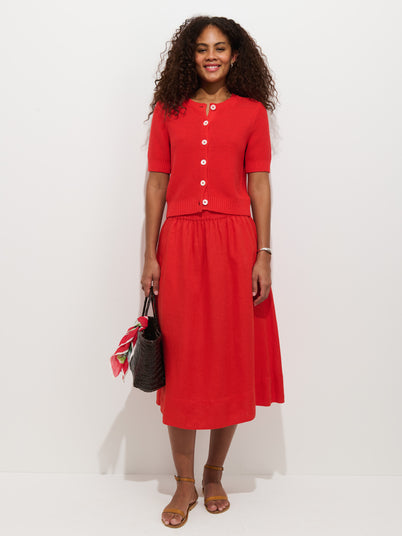 A woman with curly hair wears a bright red Maddie Skirt in Linen with a matching linen button-up top, holding a black bag with a red scarf and wearing brown sandals, standing against a plain white background.