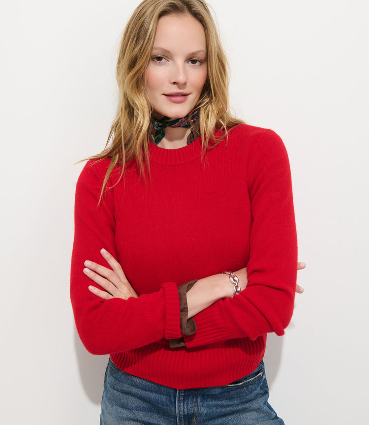 A blonde-haired woman stands against a white background, arms crossed and smiling slightly, wearing blue jeans and the Marais Sweater in Washed Cashmere—a bright red luxury sweater made from 100% cashmere—accented by a floral scarf around her neck to highlight its extra softness and elegance.
