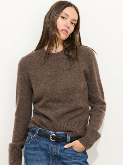 A woman with long brown hair models the Marais Crewneck Sweater in Cashmere, paired with blue jeans and a black belt, standing against a plain white background with a relaxed expression.