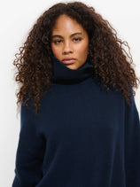 A woman with curly dark hair wears the Maisie Turtleneck Sweater in Cashmere, navy blue, relaxed fit, standing against a plain white background and looking directly at the camera with a neutral expression.