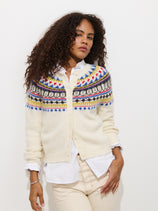 A woman with curly hair wears the Charlotte Fairisle Sweater in Wool, a cream-colored, cropped cardigan with a colorful yoke, styled over a white collared shirt and cream pants against a plain white background.