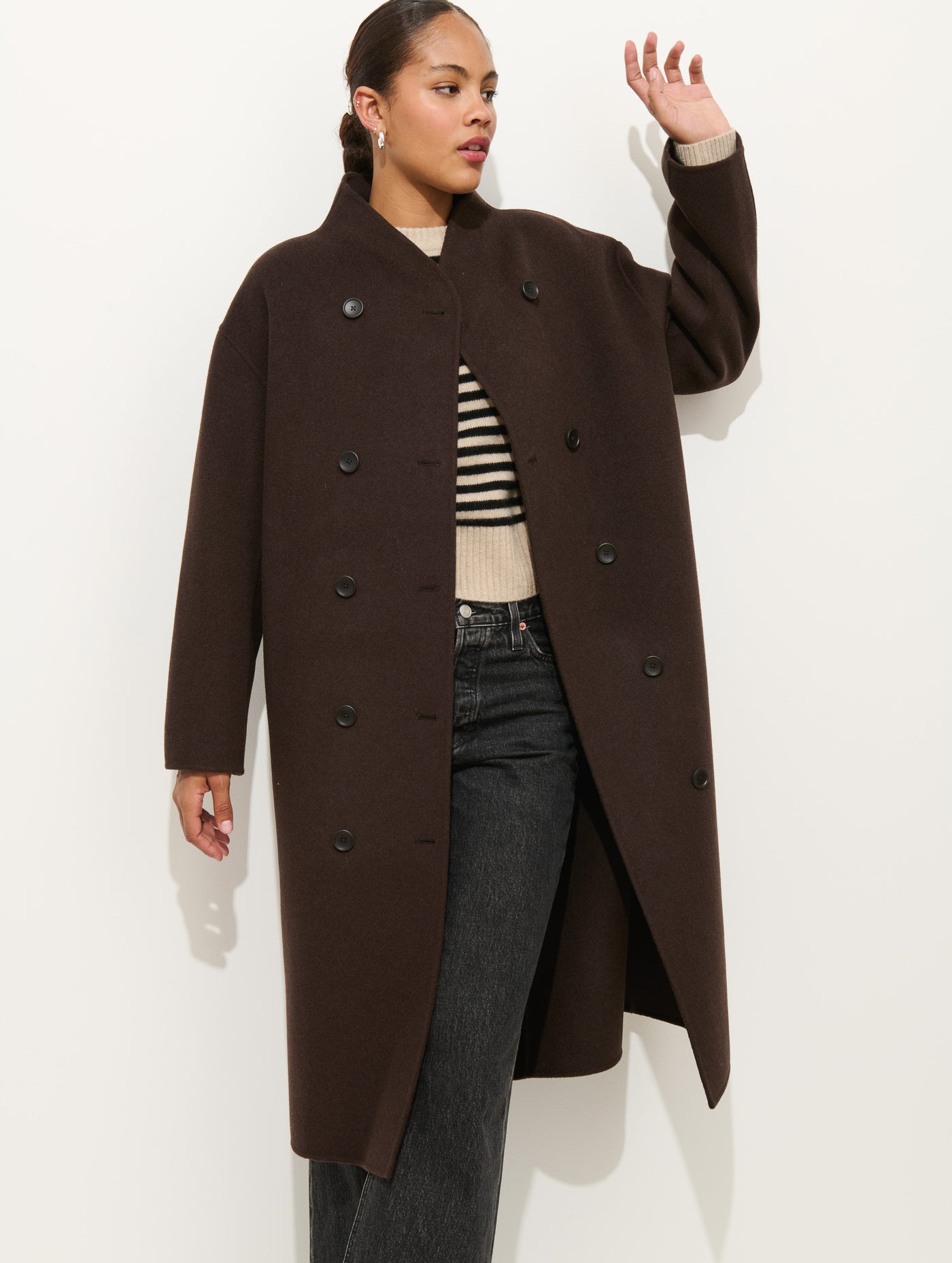 A woman stands against a white background, wearing the Long Sophia Coat— a dark brown, double-face wool coat— over a beige and black striped sweater and black jeans, with one hand raised slightly as she looks to the side.