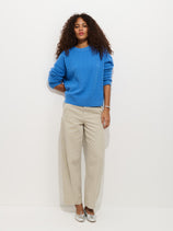 A woman with curly hair stands against a white wall in a bright blue sweater, 9th Street Pant pigment dyed wide-leg cotton pants, and silver shoes. She poses relaxedly with one hand in her pocket.