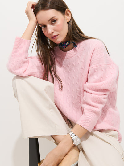 A woman in the Charlie Cable Sweater in Cashmere (light pink), beige pants, and a watch sits on a stool with one knee up. She has straight brown hair and wears a dark patterned scarf around her neck.