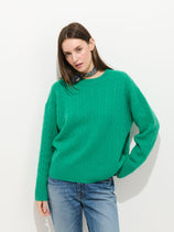 A woman in a vibrant green Charlie Cable Sweater in Cashmere, blue jeans, and a patterned scarf stands against a plain white background.