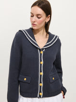 A woman models the Cassie Sailor Cardigan in cotton blend—navy blue with white trim, brass buttons, and a sailor-style collar—standing against a plain white background, looking down and slightly to the side.