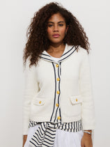 A woman with curly hair wears the Cassie Sailor Cardigan in cotton blend with brass buttons and a sailor-style collar over a white and navy striped top and skirt, standing against a plain white background.