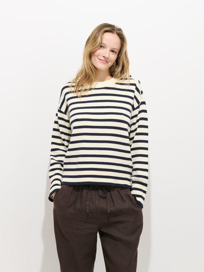 A person with shoulder-length blonde hair, smiling against a plain white background, wears the Normandy Striped Sweater in Cotton—black and white, 100% cotton, 7-gauge knit—and dark, loose-fitting pants with hands in pockets.