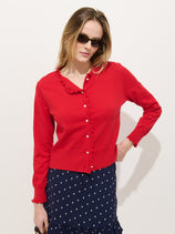 A woman in sunglasses wears the Bella Cardigan In Cotton Cashmere, a bright red sweater with white buttons, paired with a navy blue polka dot skirt, standing against a plain white background.