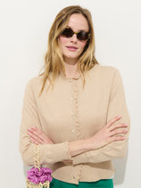 A woman with long blonde hair and sunglasses wears the Bella Cardigan in Cotton Cashmere, standing against a white background with arms crossed, holding a small woven bag featuring a purple ruffle detail.
