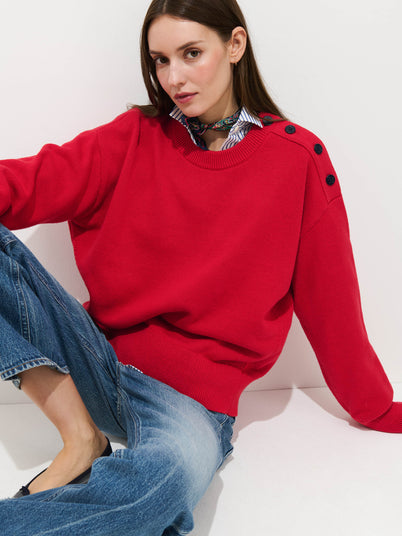 A woman with straight brown hair sits casually against a white wall, wearing the bright red Normandy Sweater in Cotton with nautical shoulder buttons, blue jeans, and a patterned neck scarf.