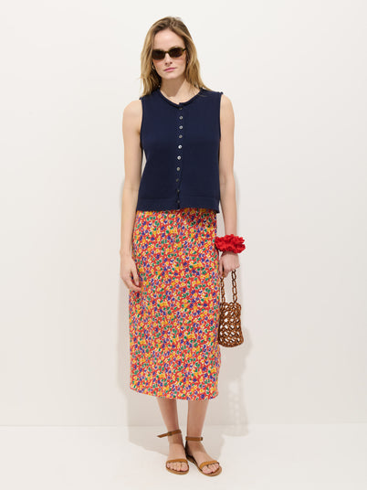A woman stands against a plain white background wearing sunglasses, a sleeveless navy buttoned top, tan sandals, holding a brown woven handbag with a red flower accessory, and the Lilly Floral Skirt—a colorful floral midi skirt with a drawcord waist.